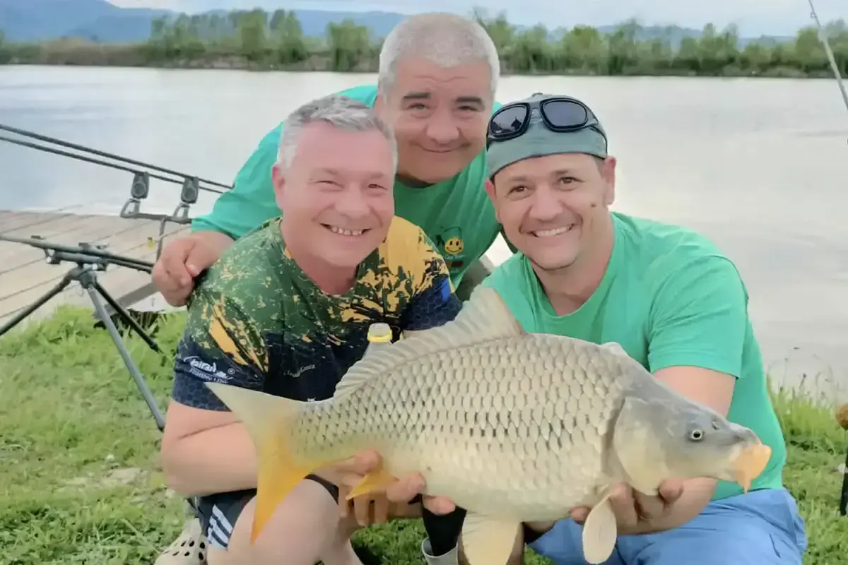 Pescari sportivi tinand un peste la C&R Lacurile Sibiu cu undite și balta de pescuit in fundal